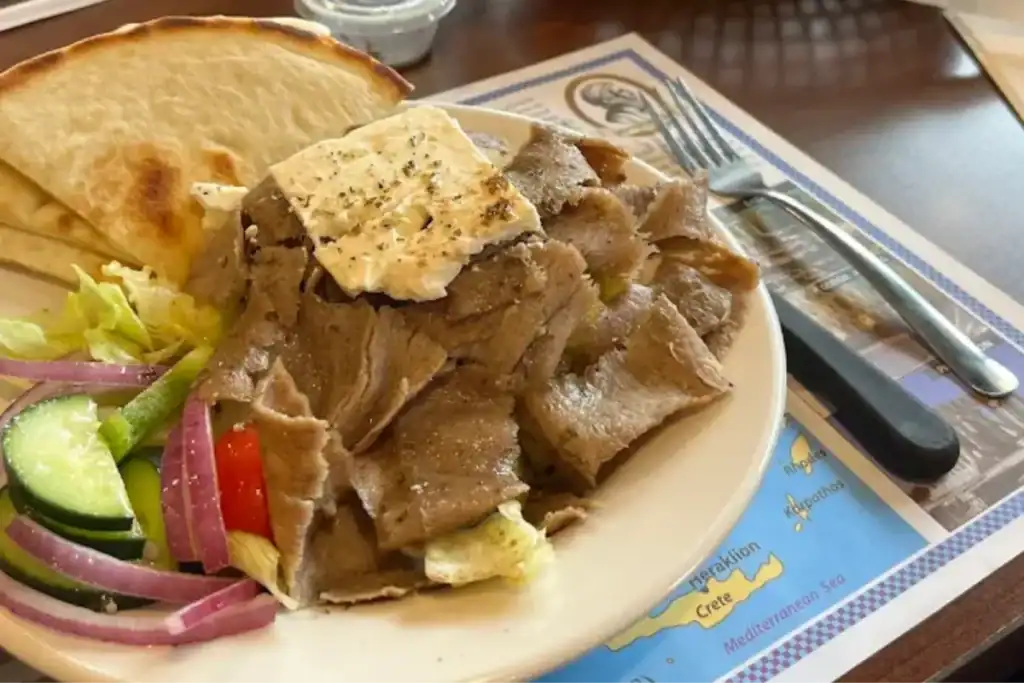 Greek restaurant gyro plate with sliced meat, pita bread, salad, and sauces.